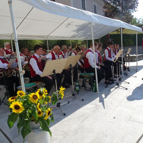 Ein Blasorchester sitzt unter einem weißen Zelt auf einer gepflasterten Fläche, mit Musikinstrumenten vor ihnen.