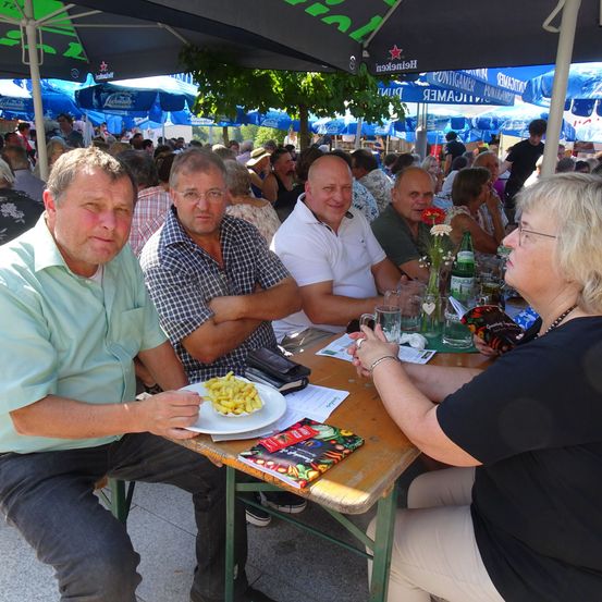 Eine Gruppe von Menschen sitzt an einem Tisch unter Schirmen bei einer Veranstaltung im Freien. Ein Mann in Grün hält einen Teller mit Pommes. Gläser, Tassen und eine Blumenvase stehen auf dem Tisch.