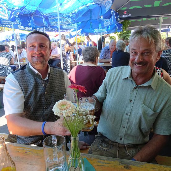 Zwei Männer sitzen an einem Tisch in einem Biergarten, lächeln und halten ein Glas Bier. Hinter ihnen sind Tische und Stühle mit sitzenden und stehenden Personen, und blaue Sonnenschirme.