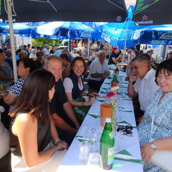 Eine Gruppe von Menschen sitzt an einem langen Tisch unter Schirmen, lächelt und unterhält sich. Der Tisch ist mit Gläsern, einer Flasche und Blumen gedeckt. Im Hintergrund stehen weitere Menschen unter Schirmen, einige stehend und unterhalten sich.