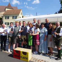 Eine Gruppe von Menschen in traditioneller Kleidung posiert auf einer Treppe für ein Foto vor einem Recycling-Behälter. Dahinter ist ein Zelt aufgestellt, und Gebäude sind im Hintergrund zu sehen.