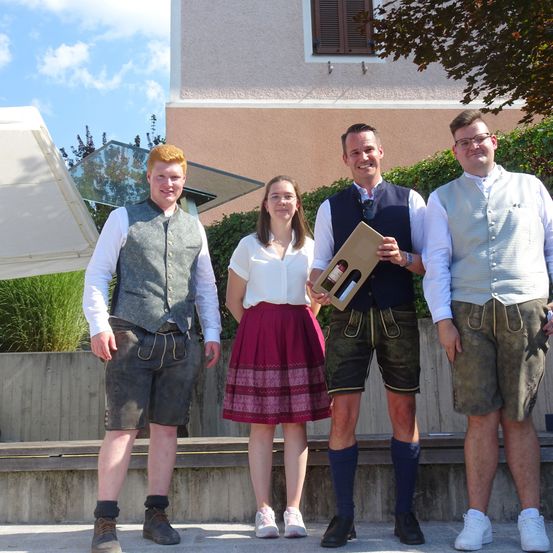 Vier Personen stehen vor einem Gebäude, zwei Männer in traditioneller bayerischer Tracht und zwei Frauen, eine im Rock und die andere im Oberteil. Sie lächeln und posieren für ein Foto.
