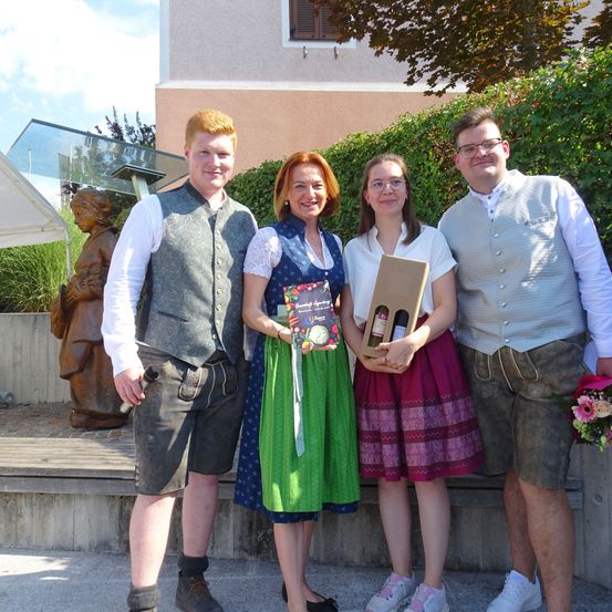 Vier Menschen in traditioneller Kleidung stehen draußen und lächeln für die Kamera. Sie halten ein Buch und eine Box. Eine Statue und Pflanzen sind hinter ihnen.