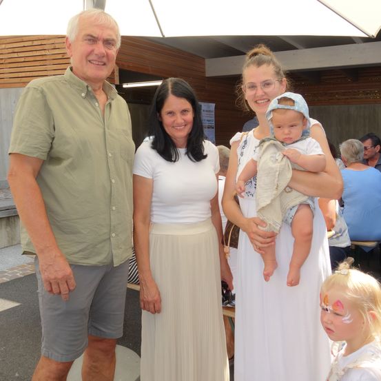 Drei Personen stehen vor einem Holzgebäude. Ein erwachsener Mann ist links, und zwei Frauen stehen neben ihm. Die Frau auf der rechten Seite hält ein Baby. Ein weiteres Kind mit Gesichtsbemalung steht neben ihr.