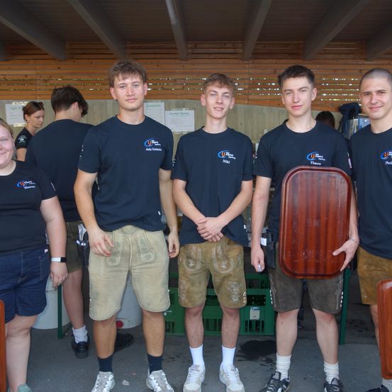 Fünf junge Menschen stehen nebeneinander vor einer Holzwand. Sie tragen blaue T-Shirts und braune Shorts. Sie lächeln für ein Foto. Dahinter stehen andere Leute und grüne Kisten.