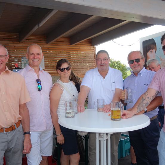 Eine Gruppe von Menschen, wahrscheinlich Freunde, stehen um einen Tisch mit Biergläsern. Sie lächeln und scheinen für ein Foto zu posieren. Hinter ihnen ist eine Holzwand.