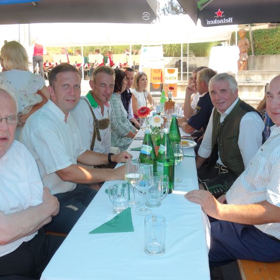 Eine Gruppe von Männern und Frauen sitzt an einem langen Tisch, lächelt und hat grüne Flaschen und Weingläser auf dem Tisch.