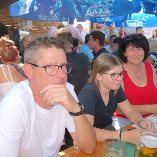 Mehrere Menschen sitzen unter blauen Sonnenschirmen um einen Tisch, darunter ein Mann mit Brille und zwei Frauen. Eine Frau hat ein Bierglas vor sich.