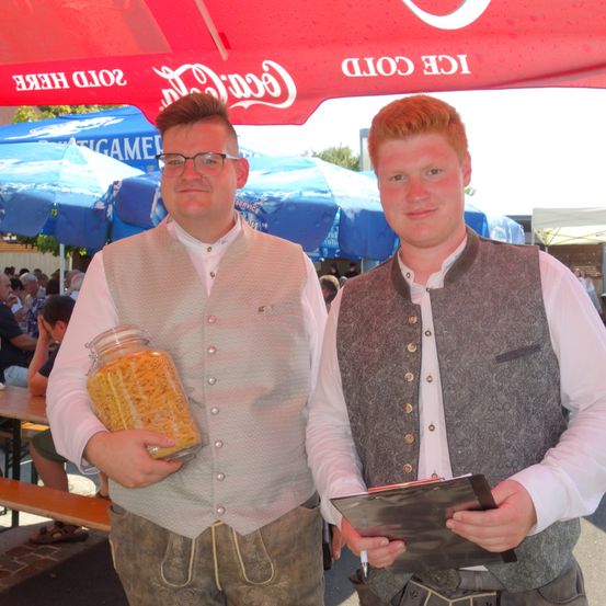 Zwei Männer stehen unter einem Coca-Cola-Zelt, einer hält eine Glaspasten und der andere eine Mappe. Sie tragen traditionelle Kleidung. Hinter ihnen sind Regenschirme und eine Menschenmenge.