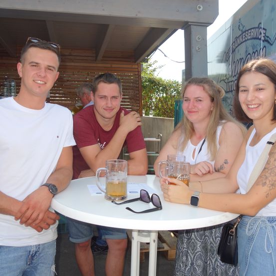 Vier Personen sitzen um einen Tisch herum, lächeln und halten Biergläser. Zwei Frauen und zwei Männer, alle in lässiger Kleidung. Dahinter steht ein Gebäude mit einer Holzwand und einem Schild.