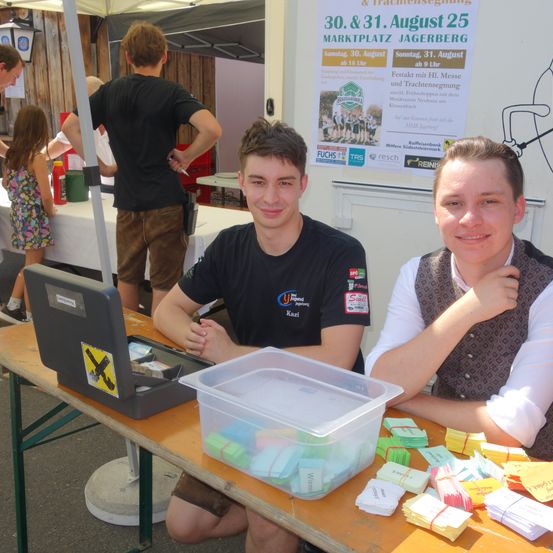 Zwei junge Männer sitzen an einem Tisch mit einer Aufbewahrungsbox und kleinen Karten. Sie lächeln und posieren für ein Foto. Hinter ihnen wirbt ein Banner für eine Veranstaltung am 30. und 31. August.