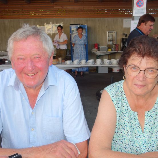 Ein älteres Paar posiert für ein Foto bei einer Veranstaltung, lächelnd, mit einem Tisch voller Tassen und Essen im Hintergrund, und einer Frau in einem blauen Kleid.