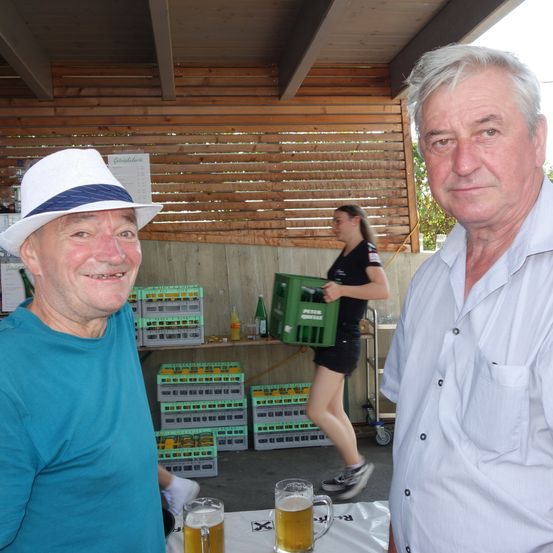 Zwei ältere Männer lächeln, einer in einem blauen T-Shirt und der andere in einem weißen Hemd. Sie haben Biergläser vor sich. Hinter ihnen ist eine Frau in Schwarz, die eine grüne Kiste trägt.