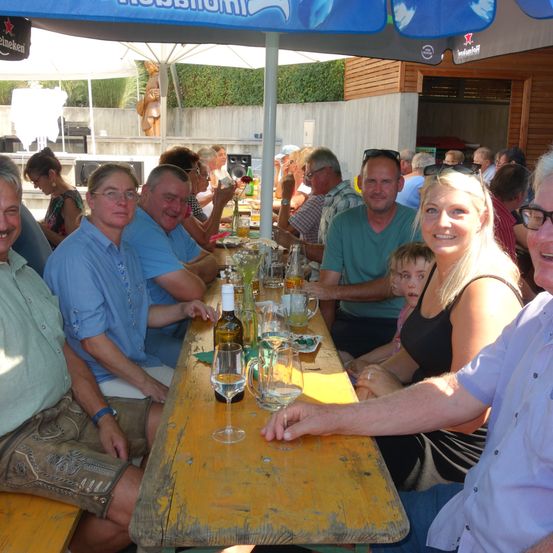 Eine Gruppe von Menschen sitzt an einem langen Tisch unter einem Schirm mit Getränken und Essen, einige tragen Brillen und Hüte.