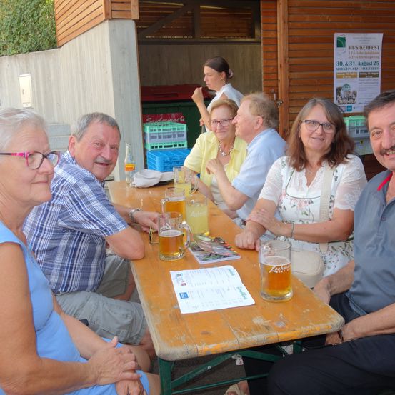 Eine Gruppe von Erwachsenen sitzt lächelnd an einem Holztisch. Es gibt Gläser mit Bier und eine Speisekarte auf dem Tisch. Eine Frau im Hintergrund schaut weg.