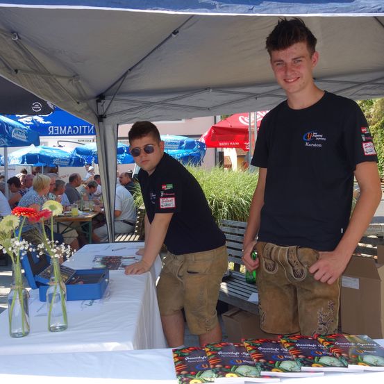 Zwei junge Männer stehen unter einem Zelt mit Büchern und Blumen auf einem Tisch, während Menschen unter blauen und roten Schirmen an Tischen sitzen.
