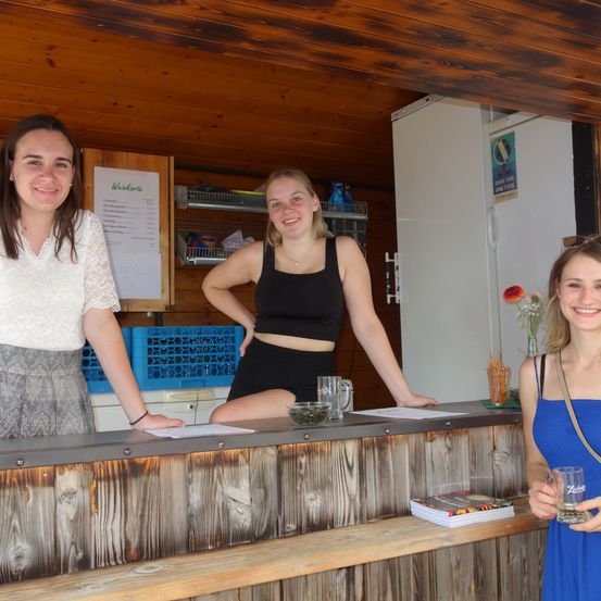 Drei Frauen stehen lächelnd hinter einer Theke. Die Frau auf der linken Seite trägt ein weißes Oberteil und eine graue Hose. Die Frau in der Mitte trägt ein schwarzes Crop-Top und schwarze Shorts. Die Frau auf der rechten Seite trägt ein blaues Kleid und hält ein Glas. Hinter ihnen befindet sich ein Kühlschrank und ein Regal.