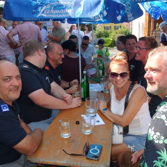Eine Gruppe von Erwachsenen sitzt unter einem Regenschirm an einem Tisch, lächelt und posiert für ein Foto. Flaschen mit Getränken und Gläser sind auf dem Tisch. Der Mann auf der linken Seite trägt eine Uhr und die Frau in der Mitte trägt eine Sonnenbrille.