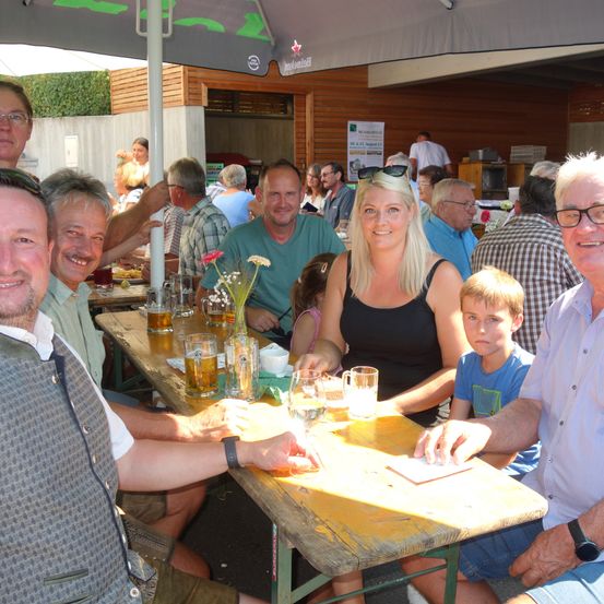 Eine Gruppe von Menschen sitzt an einem Tisch unter einem Regenschirm und lächelt für ein Foto. Sie sind von verschiedenen Getränken und Essensartikeln umgeben.