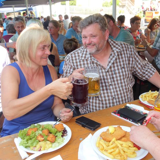 Ein Mann und eine Frau sitzen an einem Tisch, lächeln und halten Gläser mit Bier. Sie sind von anderen Menschen bei einer überfüllten Veranstaltung im Freien umgeben. Sie genießen ihre Mahlzeiten, die Salate, frittiertes Essen und Pommes Frites umfassen.