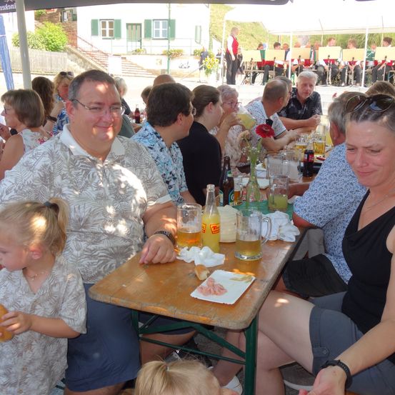 Eine Familie sitzt an einem Picknicktisch im Freien mit Getränken und Essen vor sich. Eine erwachsene Frau lächelt, während ein junges Mädchen isst. Dahinter spielt eine Band auf einer Bühne.