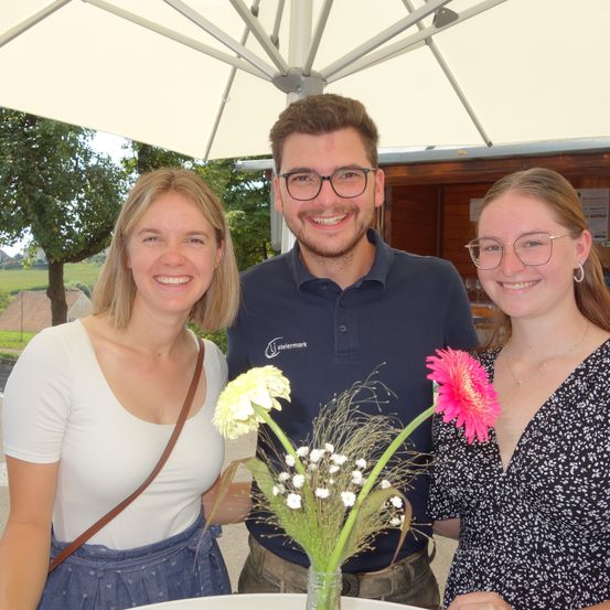 Drei Menschen stehen lächelnd unter einem Regenschirm, zwei Frauen und ein Mann. Sie halten einen Blumenstrauß. Der Mann trägt ein marineblaues Poloshirt. Die Frauen tragen weiße und schwarze Kleider.
