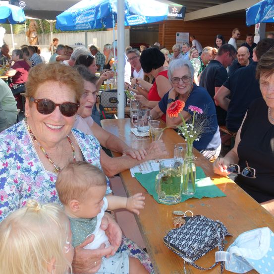 Eine Gruppe älterer Menschen, darunter ein Baby, sitzen an einem Tisch in einem Außenrestaurant. Sie lächeln und scheinen ihre Mahlzeit zu genießen.