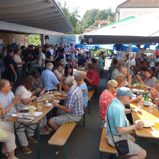 Eine große Gruppe von Menschen sitzt unter Sonnenschirmen in einem Außenbereich an Tischen. Einige essen, andere unterhalten sich, und einige Tische haben Getränke und Essen. Die Tische sind in Reihen angeordnet, und es gibt mehrere Schirme, die Schatten spenden. In der Ferne sind ein Lastwagen und einige Bäume zu sehen.