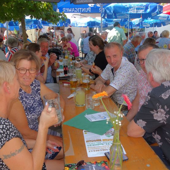 Mehrere Personen sitzen an einem langen Tisch in einer Outdoor-Einrichtung, möglicherweise einem Biergarten. Sie trinken und essen. Es gibt blaue Schirme, und ein Baum ist im Hintergrund.