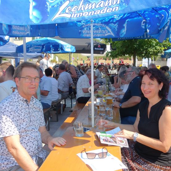 Eine Gruppe von Menschen sitzt an einem langen Tisch unter blauen Sonnenschirmen, lächelt und hält Getränke. Der Tisch ist mit Gläsern und Essen gedeckt. Die Sonnenschirme sind mit Lechman Limonaden beschriftet.