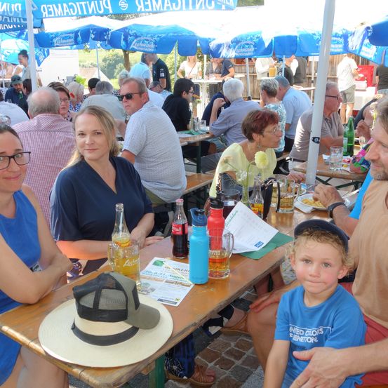 Eine Familie sitzt an einem Tisch in einem Biergarten im Freien. Sie haben Bierflaschen, eine Speisekarte und einen Hut auf dem Tisch. Um sie herum sitzen Menschen an Tischen unter Schirmen. Die Atmosphäre ist locker und lebhaft.