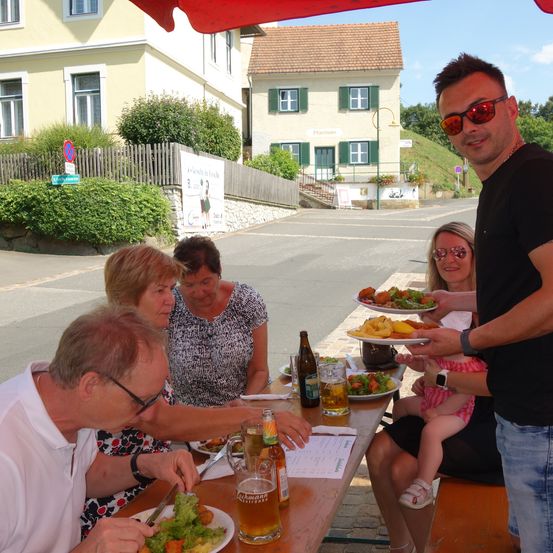Eine Familie isst im Freien an einem Picknicktisch, wobei ein Mann ein Baby hält. Der Tisch ist mit Tellern voller Essen und Bier gedeckt. Dahinter ist ein Haus mit einer Straße und einem Hügel zu sehen.