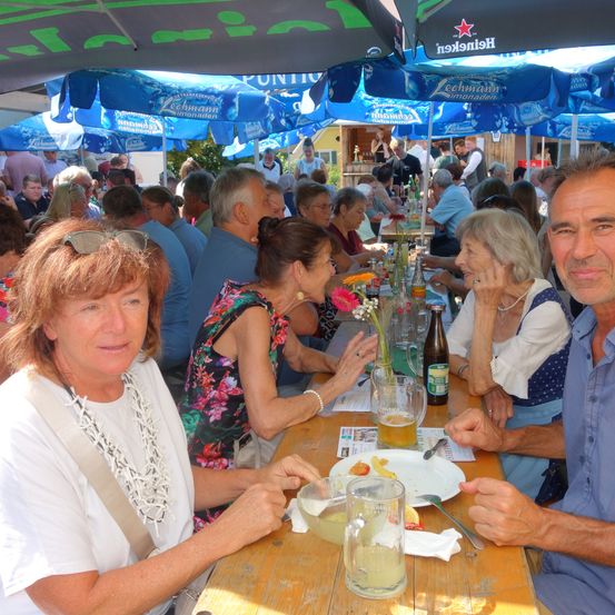 Eine Gruppe von Menschen sitzt an einem langen Tisch unter blauen Sonnenschirmen und trinkt Bier und isst Essen.
