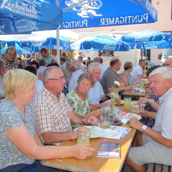 Eine Gruppe von Menschen sitzt an Tischen unter blauen Sonnenschirmen. Sie genießen Essen und Getränke. Ein Mann trägt eine Brille und eine Uhr. Die Sonnenschirme haben Text darauf.