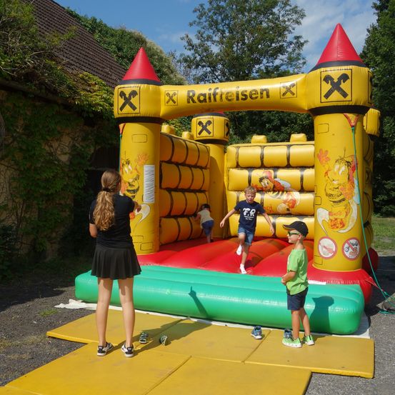 Eine Gruppe von Kindern spielt in einem großen gelben und roten aufblasbaren Sprunghaus. Ein Kind springt hinein, während andere zuschauen und eine Frau in der Nähe steht.