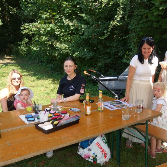 Eine Gruppe von Menschen versammelt sich um einen Tisch im Freien. Eine Frau malt, während andere zusehen. Auf dem Tisch stehen Flaschen, Gläser und eine Schale mit Farbe. Ein Kinderwagen steht in der Nähe, und ein Kind steht neben einer Bank. Bäume sind im Hintergrund.