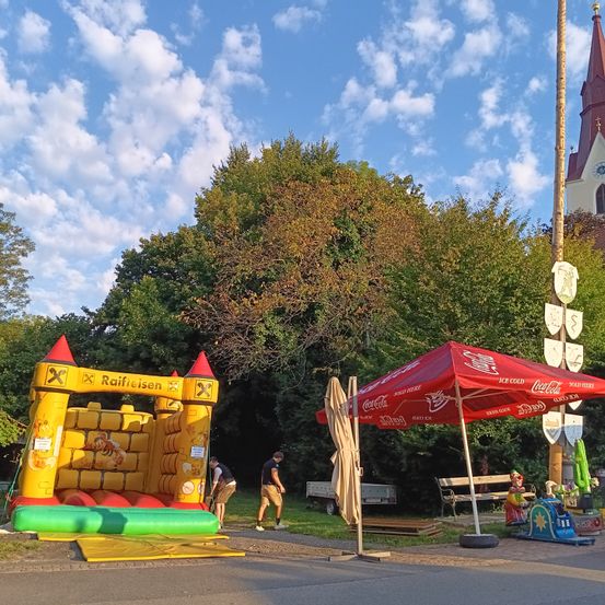 Ein aufblasbares Schloss mit Raiffeisen-Marke und einem Coca-Cola-Schirm ist draußen aufgebaut, wobei zwei Personen es aufbauen und eine Kirche im Hintergrund steht.