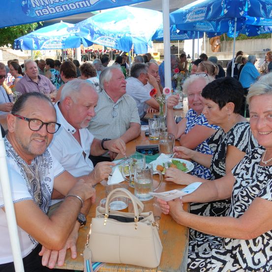 Eine Gruppe von Menschen sitzt an einem Außentisch unter blauen Sonnenschirmen. Sie essen und trinken. Eine Frau rechts lächelt. Eine Handtasche steht auf dem Tisch.
