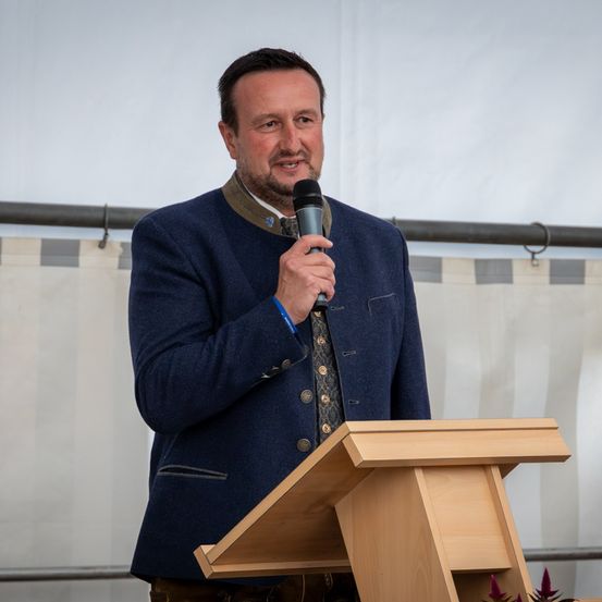 Ein Mann steht an einem Podium, hält ein Mikrofon in der Hand und scheint eine Rede zu halten. Er trägt einen blauen Anzug und eine dekorative Weste. Hinter ihm befindet sich ein weißer Hintergrund.