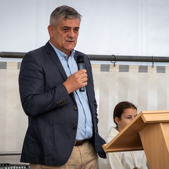Ein Mann in einem blauen Hemd und Blazer spricht in ein Mikrofon am Rednerpult, während ein junges Mädchen im Hintergrund zuhört.