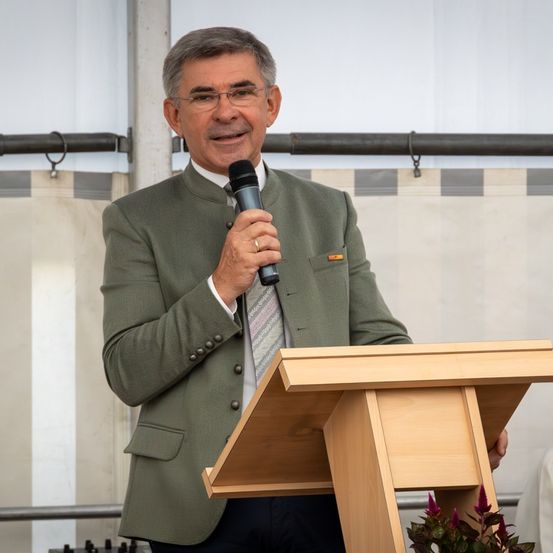 Ein Mann in einer grünen Jacke und Krawatte steht an einem Podium, hält ein Mikrofon in der Hand und lächelt. Er scheint eine Rede oder Präsentation zu halten.