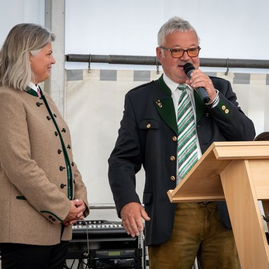 Ein erwachsener Mann spricht in ein Mikrofon, während er an einem Rednerpult steht. Eine erwachsene Frau steht neben ihm und hört zu.