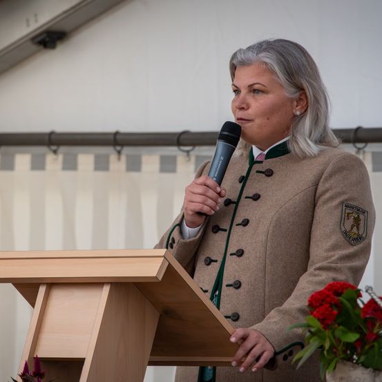 Eine Frau steht an einem Rednerpult mit Mikrofon und hält eine Rede. Sie trägt einen braunen Mantel mit grünem Besatz und einem Abzeichen. Hinter ihr ist eine weiße Wand zu sehen.