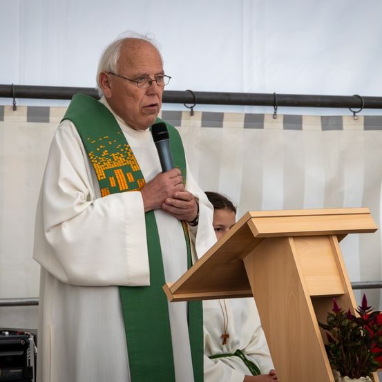 Ein älterer Priester in einer weißen Robe und einem grünen Stola spricht in ein Mikrofon an einer Kanzel, mit einem Kind in einer weißen Robe, das hinter ihm sitzt, und einer Pflanze in einem Blumentopf rechts.