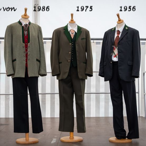 Drei Mannequins präsentieren Herrenanzüge aus verschiedenen Jahren: 1956, 1975 und 1986. Jeder Anzug ist mit dem Jahr beschriftet und zeigt Unterschiede in Stil und Stoff.