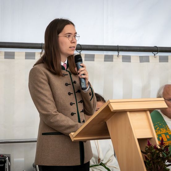 Eine Frau mit Brille spricht in ein Mikrofon an einem Podium, sie trägt einen braunen Mantel mit grünem Besatz. Hinter ihr sitzt ein Mann in einem grünen Gewand.