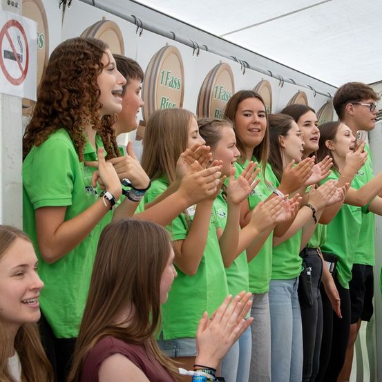 Eine Gruppe junger Leute in grünen T-Shirts steht in einer Reihe, klatscht und lächelt, während einer ein Foto macht. Sie befinden sich in einem weißen Zelt mit Fässern und einem Rauchverbotsschild.