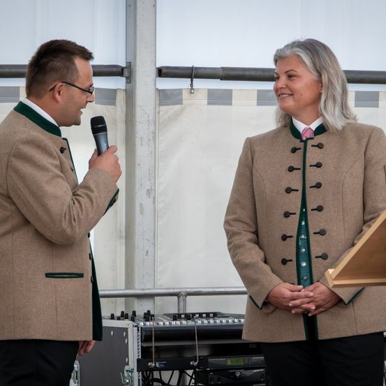 Ein Mann in einem beigen Mantel, der ein Mikrofon hält, spricht zu einer Frau in einem beigen Mantel mit grünen Akzenten und einer rosa Krawatte.