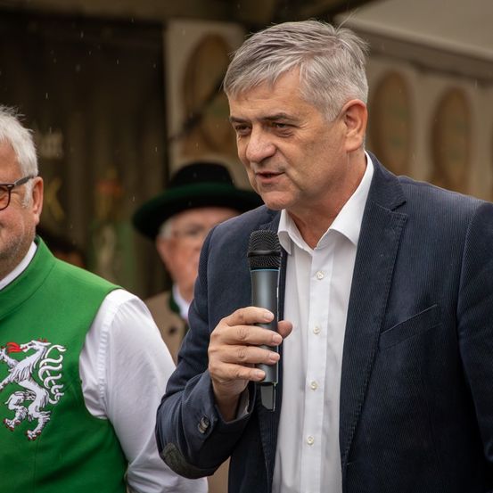 Ein Mann in einem Anzug hält ein Mikrofon und spricht bei einer Veranstaltung im Freien, während ein anderer Mann in traditioneller Kleidung hinter ihm steht.