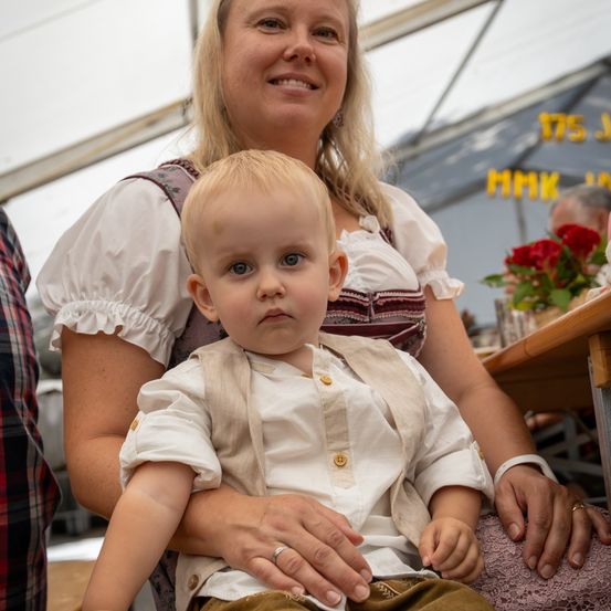 Eine Frau mit blonden Haaren in einem traditionellen Kleid hält ein Baby in einem weißen Hemd und einem beigen Westchen. Sie sitzen in einem Zelt mit lächelndem Gesicht.